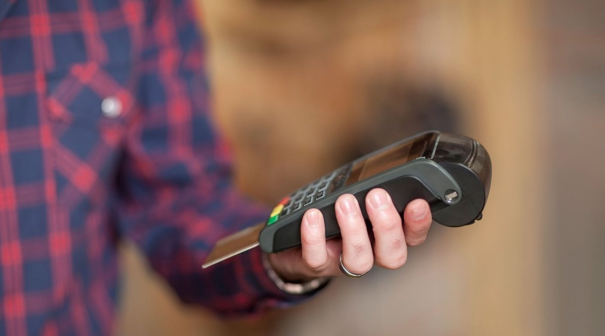 A person wearing a blue and red checked shirt holds a card reader waiting for a transaction to go through